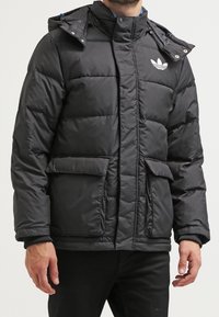 Svart pufferjacka med huva, dragkedja, två framfickor och vitt logo på bröstet. Vadderad design med en slät yta.