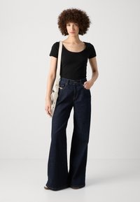 Černé tričko s výstřihem do lodě, obepínající postavu, spárované s vysokými širokými džíny z tmavého denimu. Na těle modelky odpočívá světle zbarvená kabelka přes rameno.