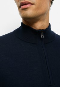 Marineblauer Reißverschluss-Pullover mit hohem Kragen, gerippter Struktur und Reißverschluss am Hals, mit glatter Oberfläche und minimalistischem Design.
