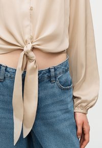 Μπεζ σατέν μπλούζα με κουμπιά, δέσιμο μπροστά σε κόμπο, σε συνδυασμό με τζιν ψηλόμεσα μπλε, που αφήνει μέρος της γυμνής μέσης εμφανές.