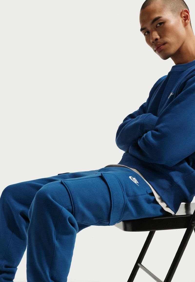 Sweat-shirt et pantalon Nike bleus en tissu doux, avec un logo et plusieurs poches. Texture lisse avec une coupe décontractée.