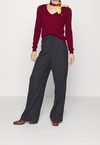 Burgunderfarbene V-Ausschnitt-Strickjacke kombiniert mit hoch taillierten, weit geschnittenen schwarzen Hose. Braune, lackierte Lederschuhe und ein Seidenschal mit gelben Akzenten.