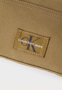 Rudas audinio pleistras su tekstu "Calvin Klein Jeans" ir juodu „CK“ logotipu, apvaduojamas baltomis siūlėmis. Tekstūruotas fonas.