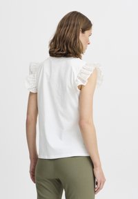 Top en coton blanc avec des manches volantes en dentelle à œillets ; texture douce ; coupe décontractée. Associé à un pantalon vert olive, avec un branding minimal visible.