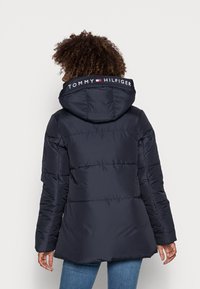 Marineblå polstret jakke med hætte. Har vandret syninger, logo på kraven og en let flared kant. Stoffet fremstår glat og glansfuldt.