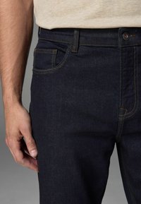 Los jeans de mezclilla azul oscuro presentan un diseño clásico de cinco bolsillos con costuras naranjas en contraste y un cierre de botón metálico. Textura suave.