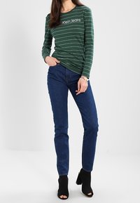 Grünes, langärmliges gestreiftes Hemd mit weißen Linien, versehen mit dem Logo "Klein Jeans". Tragen mit eng anliegenden dunkelblauen Jeans und schwarzen offenen Stiefeletten.