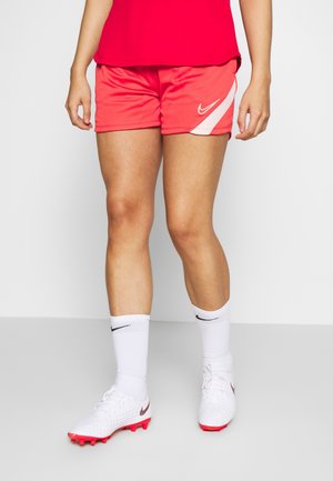 Person iført røde sportsshorts, hvide Nike-strømper og hvide fodboldstøvler med røde såler, stående på en flad overflade.