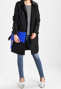 Black trench coat with double-breasted design, paired with a striped shirt, light denim jeans, silver flats, and a blue clutch bag.