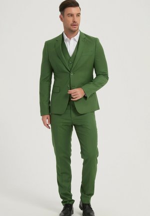 Homme portant un costume trois pièces vert et une chemise blanche, debout avec une main près du bouton de la veste, regardant légèrement sur le côté.