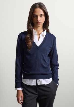 Giovane donna con i capelli lunghi e castani che indossa un maglione a V blu navy sopra una camicia bianca e pantaloni scuri, in piedi con una mano in tasca.