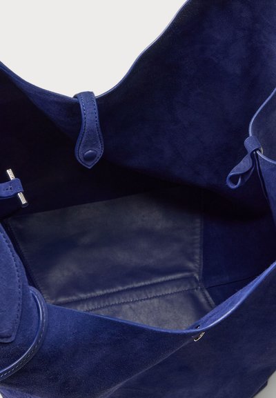 Sac fourre-tout en daim bleu marine avec un dessus ouvert, présentant un intérieur lisse, deux sangles latérales et un matériel minimal pour une fermeture sécurisée.