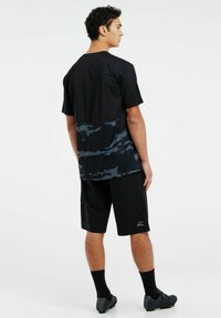 Musta urheilu t-paita, jossa on teksturoidtu, kuvioitu alaosa, ja mustat shortsit. Malli, jolla on mustat sukat ja nappulakengät, näkyy takaa.