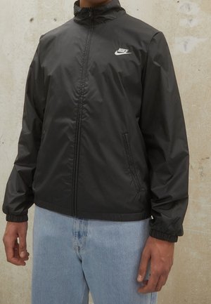 Homme portant une veste coupe-vent Nike noire zippée, associée à un jean bleu clair, debout contre un mur texturé beige.