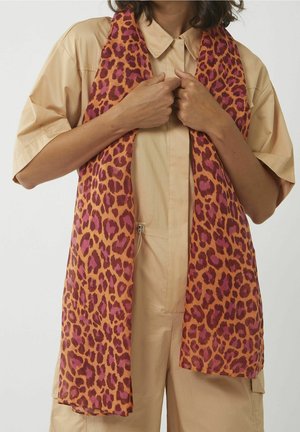 Femme portant une combinaison beige à manches courtes boutonnée avec une longue écharpe imprimé léopard orange et rose drapée autour du cou.