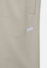 Beige Sweatpants mit einer glatten Textur, die ein kleines weißes gesticktes Logo am linken Oberschenkel und seitliche Taschen aufweisen.