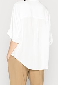 Blusa bianca, oversize, con maniche corte arrotolate, tessuto leggero, orlo curvo e dettaglio sulla parte posteriore, abbinata a pantaloni color cammello.