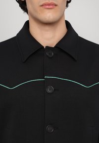 Černá košile s límečkem, vyznačující se tyrkysovým prošíváním podél hrudníku a dvěma černými knoflíky.