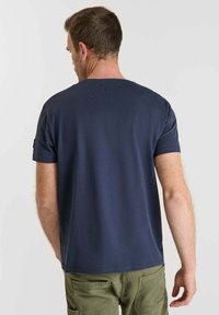 Hombre con cabello corto castaño, vistiendo una camiseta de manga corta azul marino lisa y pantalones verde olivo, visto desde atrás.