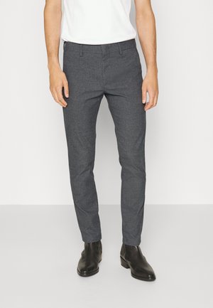 Hombre con pantalones de vestir grises ajustados, botas negras de cuero hasta el tobillo y una camisa blanca, de pie frente a un fondo liso.