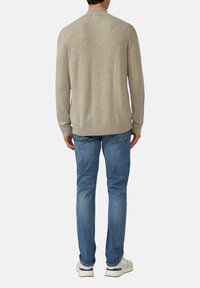 Beiger Strickpullover mit hohem Kragen, gerippten Bündchen und Saum, kombiniert mit blauen Jeans und weißen Turnschuhen. Dezente Textur sichtbar.