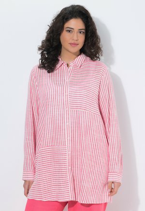 Frau mit lockigem dunklem Haar, die ein langärmeliges, rosa-weiß vertikal gestreiftes Hemd mit Knöpfen und eine rosa Hose trägt.