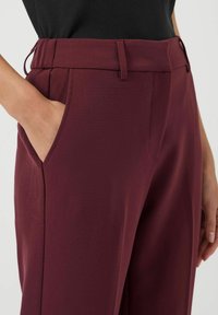 Osoba ubrana w wysokie bordowe spodnie z ręką w bocznej kieszeni, w połączeniu z czarnym topem.