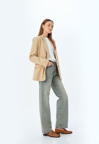Blazer beige con hombros estructurados sobre una camisa blanca, combinado con jeans de mezclilla claros de cintura alta y mocasines marrones. Accesorios sencillos.