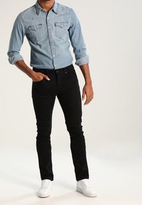 Man med ljusblå denimskjorta, svarta slim-fit jeans, vita sneakers, stående med ena handen i fickan mot en neutral bakgrund.