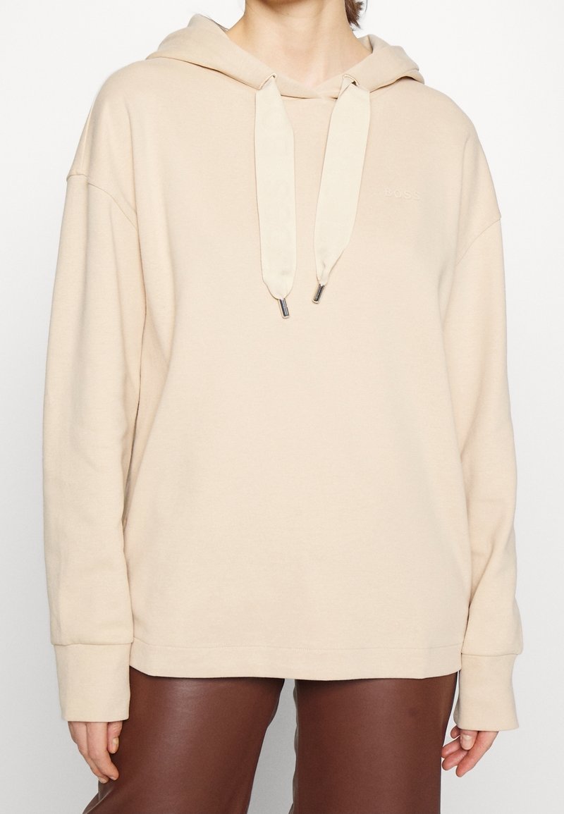 Personne portant un sweat à capuche beige avec de larges cordons et un pantalon en cuir marron, vue des épaules jusqu'au milieu des cuisses sur un fond blanc.