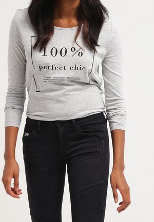 Femme portant un t-shirt à manches longues gris clair avec le texte "100% perfect chic" et un pantalon noir ajusté avec des détails côtelés au niveau des genoux.