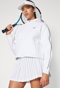 Hvit zip-up jakke med grønne detaljer, parret med et plissert hvitt skjørt. Modellen holder en tennisracket og har på seg en hvit visir.