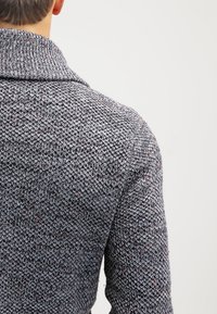 Cardigan lavorato a maglia di un grigio scuro con una trama spessa, caratterizzato da un colletto a scialle e macchie di rosso e blu in tutto il capo.