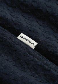 Mørkeblått teksturert stoff med en hvit rektangulær etikett sydd på, som viser merkenavnet "GABBA" i svarte store bokstaver.