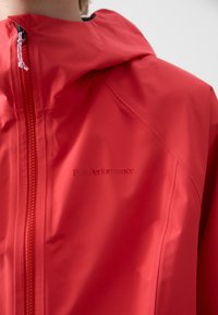 Red waterproof jacket featuring a zip closure, adjustable hood, and embossed logo on the chest. Smooth texture with a sleek design.