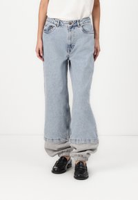 Šviesiai mėlyni džinsai su plačiomis kojomis ir aukštu liemeniu, su priekinėmis kišenėmis, sukritais kraštais, po kuriais matosi pilki treningai, ir juodi batai.