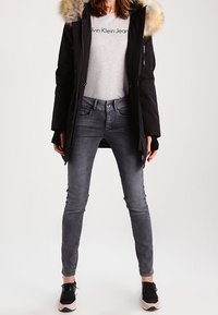 Parka negra con capucha forrada de piel, camiseta gris y jeans grises. Accesorios con zapatillas negras. Diseño simple, forma ajustada.