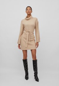 Robe beige en tricot avec un col drapé, une taille ceinturée et des manches longues, associée à des bottes noires jusqu'aux genoux. Texture lisse et design ajusté.