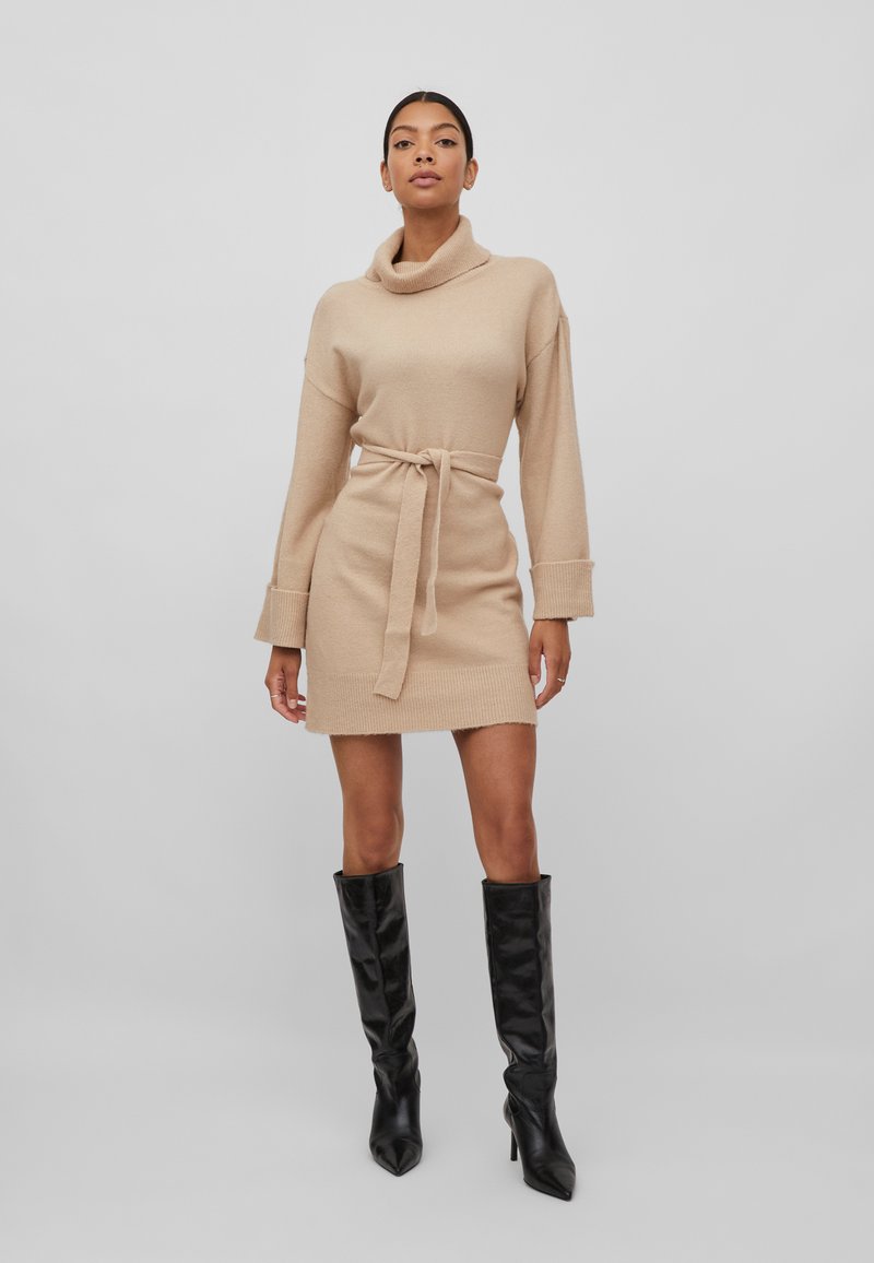 Robe beige en tricot avec un col drapé, une taille ceinturée et des manches longues, associée à des bottes noires jusqu'aux genoux. Texture lisse et design ajusté.