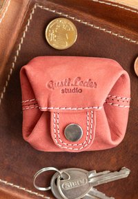 Červená kožená peněženka na mince se západkou, vybavená bílým prošíváním a embosovaným logem. Umístěná na hnědé kožené ploše vedle mincí a klíčů.