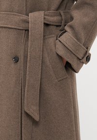 Brūns vilnas mētelis ar saistītu jostu, pogām pie piedurknēm un sānu kabatām. Audumam ir teksturēta apdare un piegriezums.