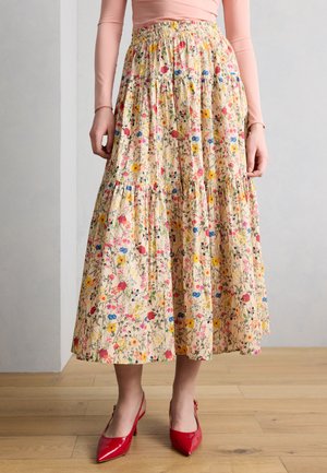 Frau trägt einen hoch taillierten, beigen Midirock mit Blumenmuster, kombiniert mit einem hellrosa Langarmshirt und roten spitzen Absätzen.