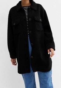 Abrigo negro de textura esponjosa con cuello tipo camisa, cierre frontal de botones y dos bolsillos en el pecho, llevado sobre una camiseta a rayas y jeans de mezclilla.