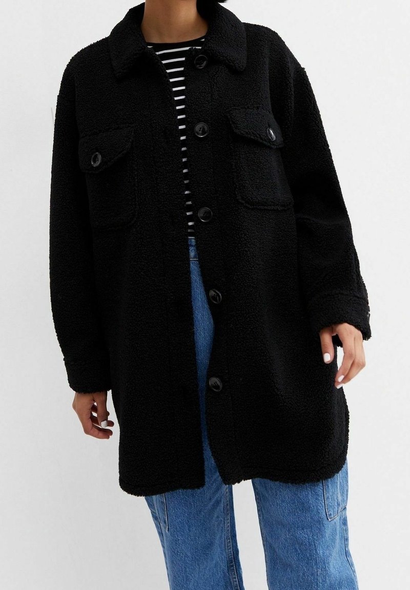 Abrigo negro de textura esponjosa con cuello tipo camisa, cierre frontal de botones y dos bolsillos en el pecho, llevado sobre una camiseta a rayas y jeans de mezclilla.