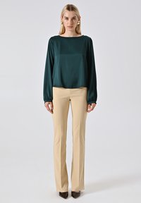 Groene satijnen blouse met lange mouwen en een wijde halslijn, gecombineerd met beige aansluitende broek, gedragen met puntige zwarte hakken.