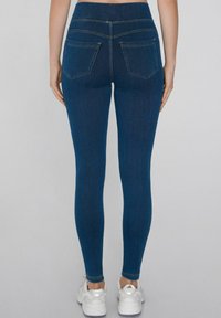 Mujer con jeans skinny de color azul oscuro y zapatillas blancas, de pie con los brazos relajados a los lados, vista desde atrás.