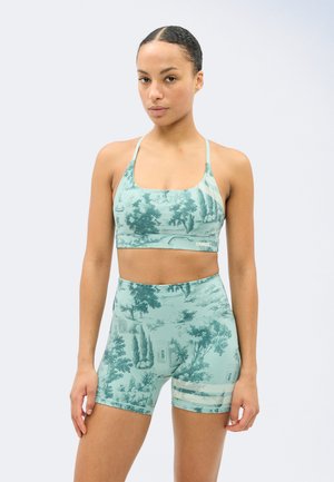 Γυναίκα στέκεται φορώντας ταιριαστό πράσινο αθλητικό σουτιέν και σορτς με ψηλή μέση, με σχέδιο φύσης και αρχιτεκτονικής σε ανοιχτό φόντο.