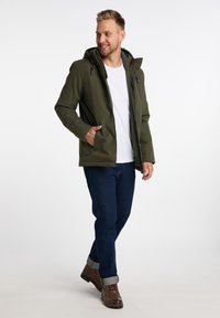 Olivgrüne Jacke mit Kapuze, Reißverschluss und seitlichen Taschen, kombiniert mit dunkelblauen Jeans und braunen Stiefeln. Weißes T-Shirt ist darunter sichtbar.