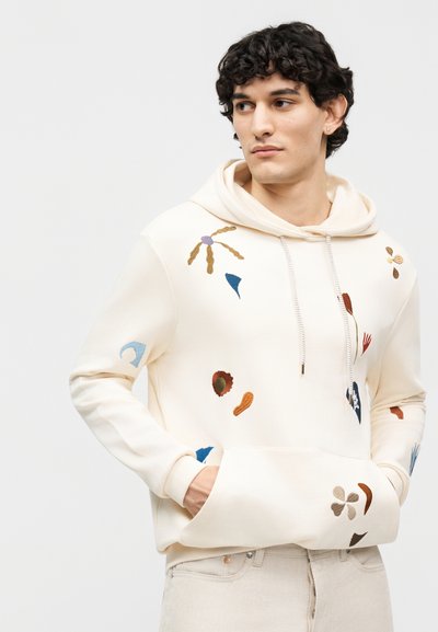 Mladý muž s kučeravými vlasmi, oblečený v smotanovej mikine s viacfarebnými abstraktnými vyšívanými tvarmi, ruky vo vrecku vpredu, hľadí do strany.