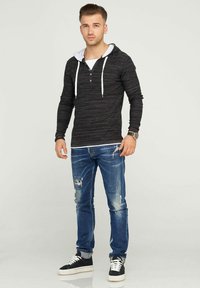 Schwarzer Hoodie mit langen Ärmeln und weißen Akzenten, über einem weißen T-Shirt getragen, kombiniert mit zerrissenen blauen Jeans und schwarzen Turnschuhen.